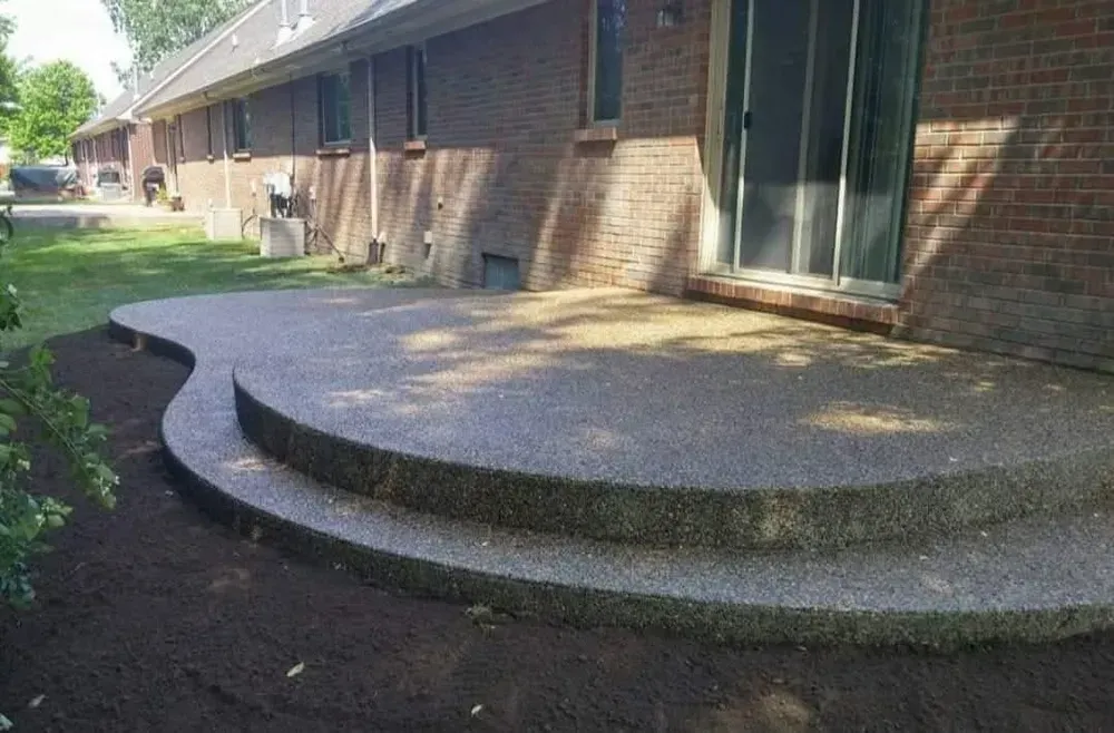 Concrete patio with steps, brick house. Dark brown soil, green grass.