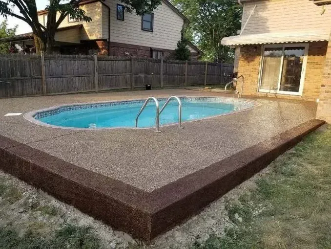 Oval pool surrounded by pebble-covered concrete patio with brown border; a house and fence are in the background.