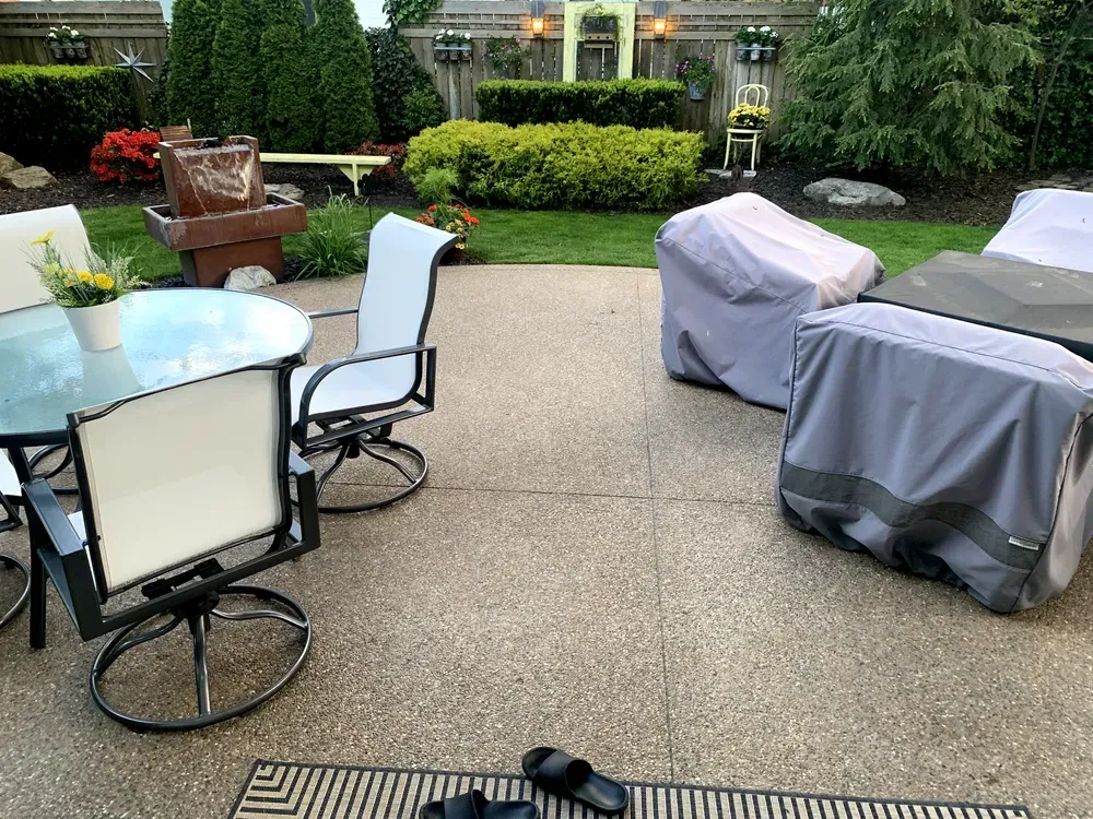 Patio with furniture, some covered with grey fabric, and a garden in the background.