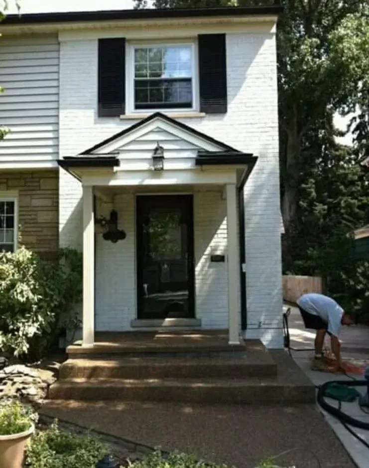 A two-story white brick house with a black door, shutters, and a person sweeping the porch.