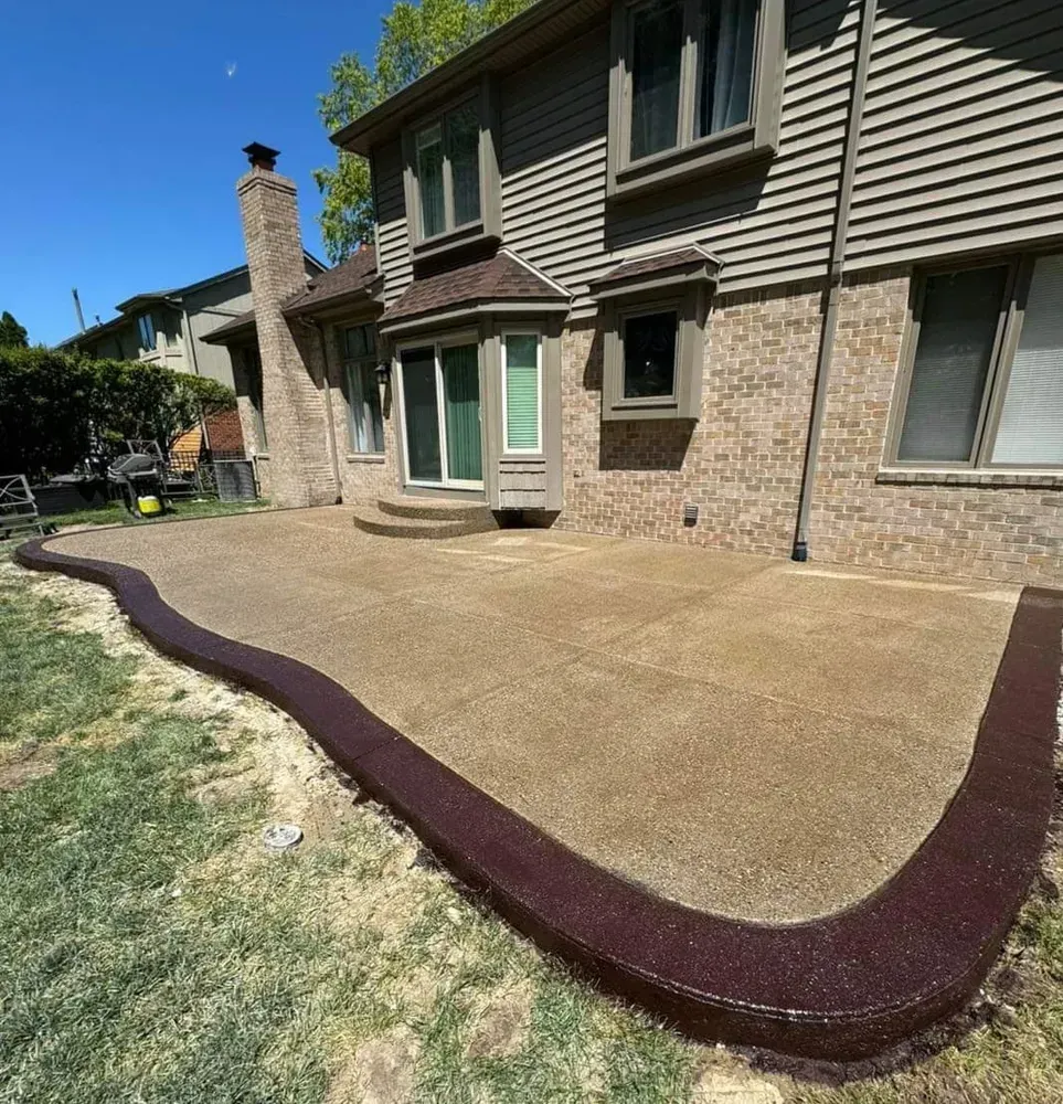 Backyard patio with a brown border, next to a two-story brick house and green lawn.