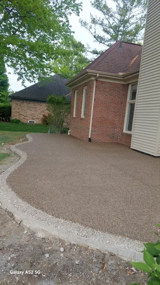 Gravel patio next to a brick house, with a concrete border. Green trees and cloudy sky in the background.