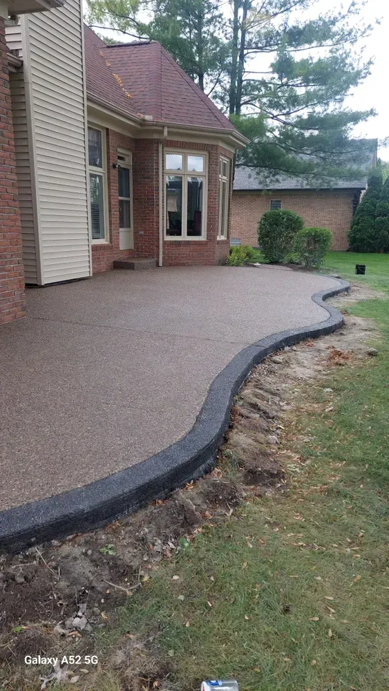 Brown concrete patio with curved black border next to a house and grassy yard.