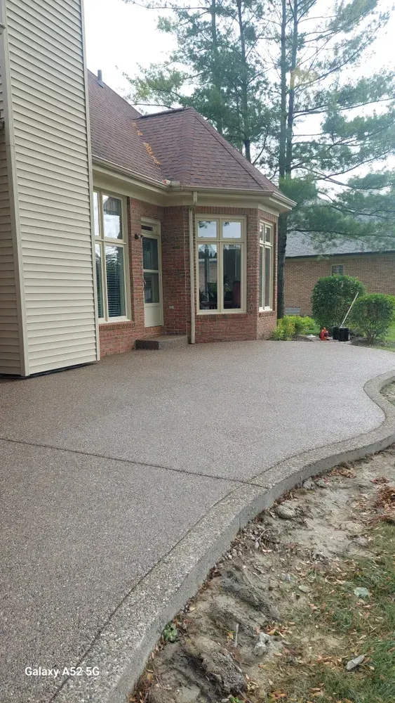 Concrete patio next to a brick house, edged with a low concrete border, overgrown grass.