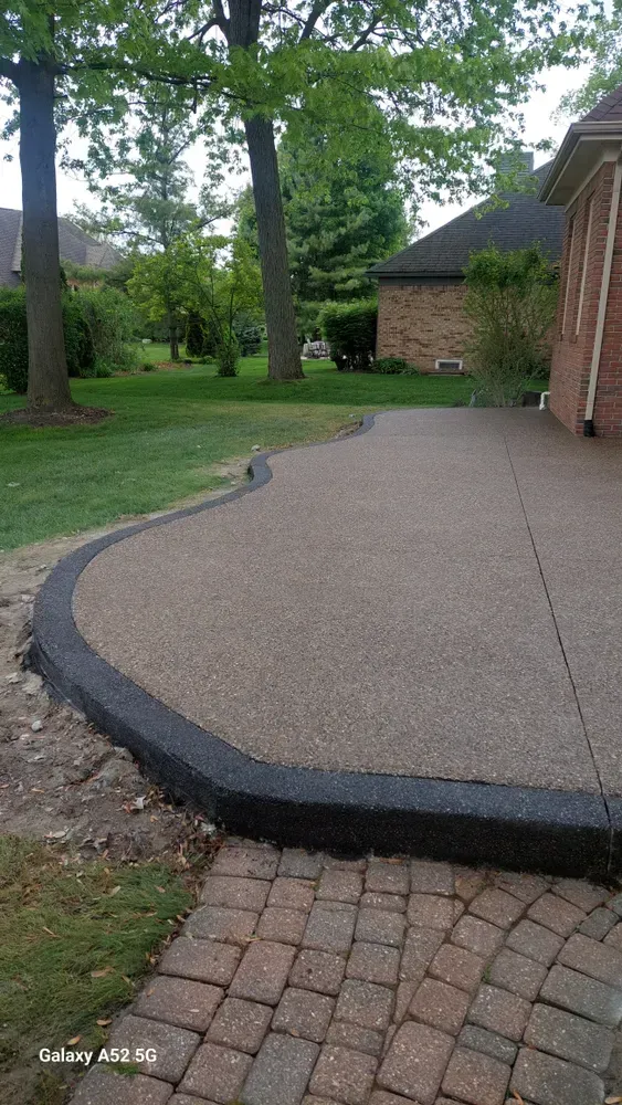 Concrete patio with dark border, curving to join lawn. Next to brick pathway, a house in the background.