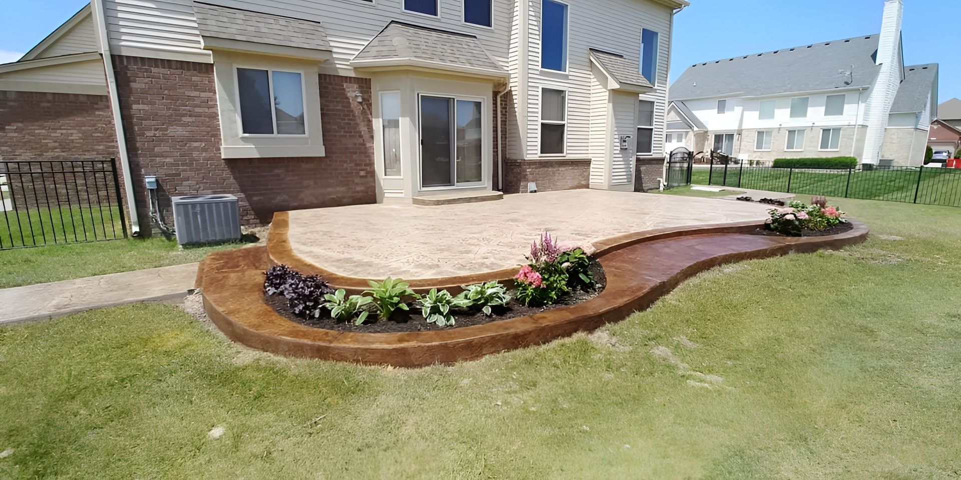Backyard patio with a flower bed border. House with a brick facade and a manicured lawn.