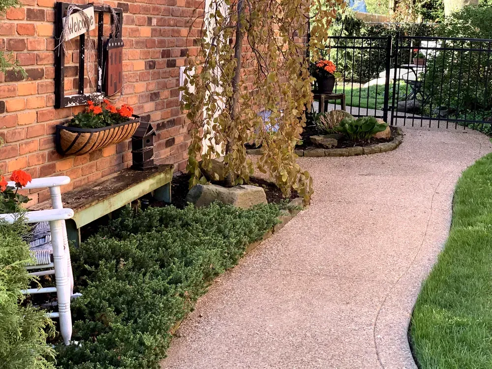 A curved pathway of stone leads through a garden next to a brick building with plants and a bench.