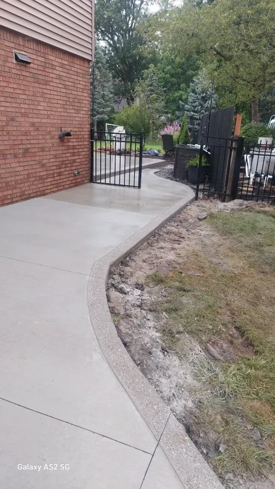 Concrete pathway with decorative edge curves towards a black gate near a brick building and landscaping.