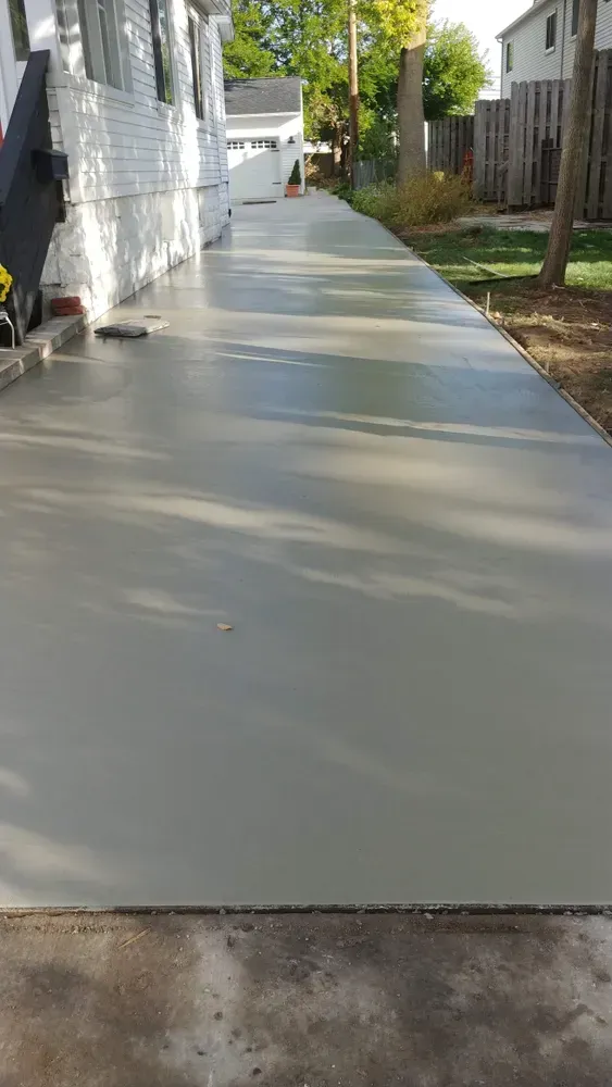 Freshly poured gray concrete sidewalk alongside a white house; a driveway leads to a garage.