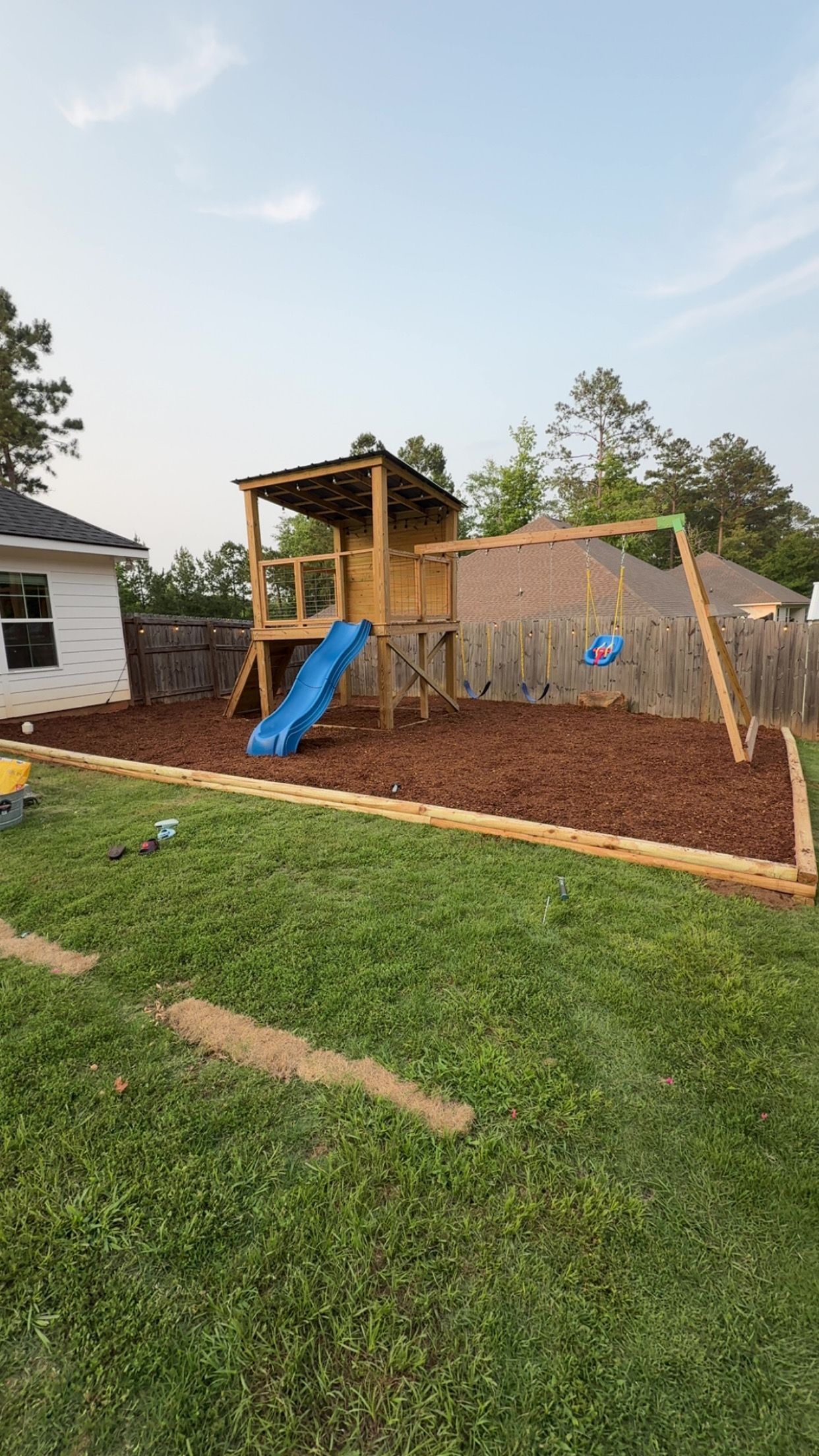 Wooden playset with slide and swing set in a backyard with wood mulch and green grass.