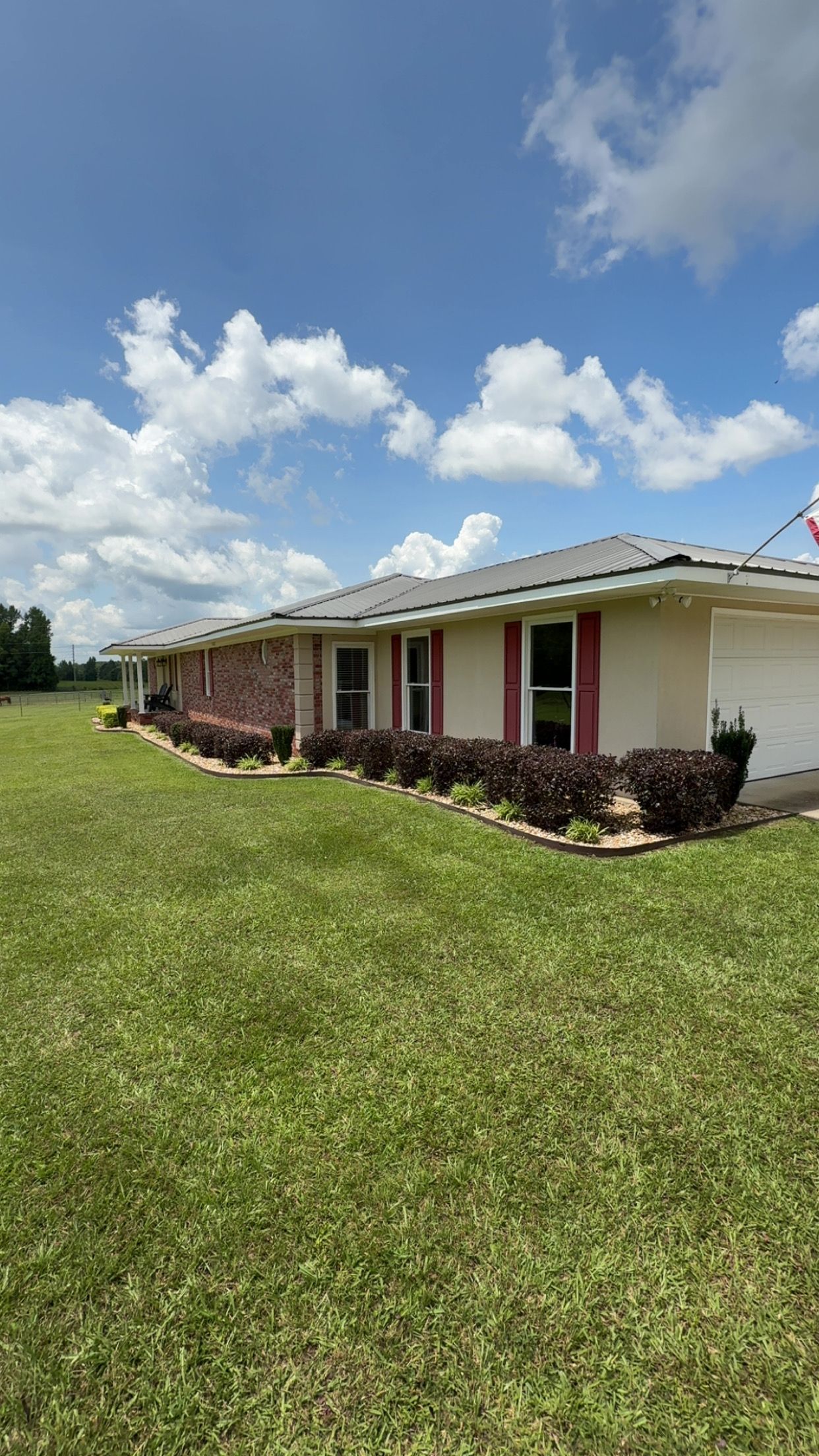 A one-story beige house with a brick accent, red trim, and a green lawn under a partly cloudy sky.