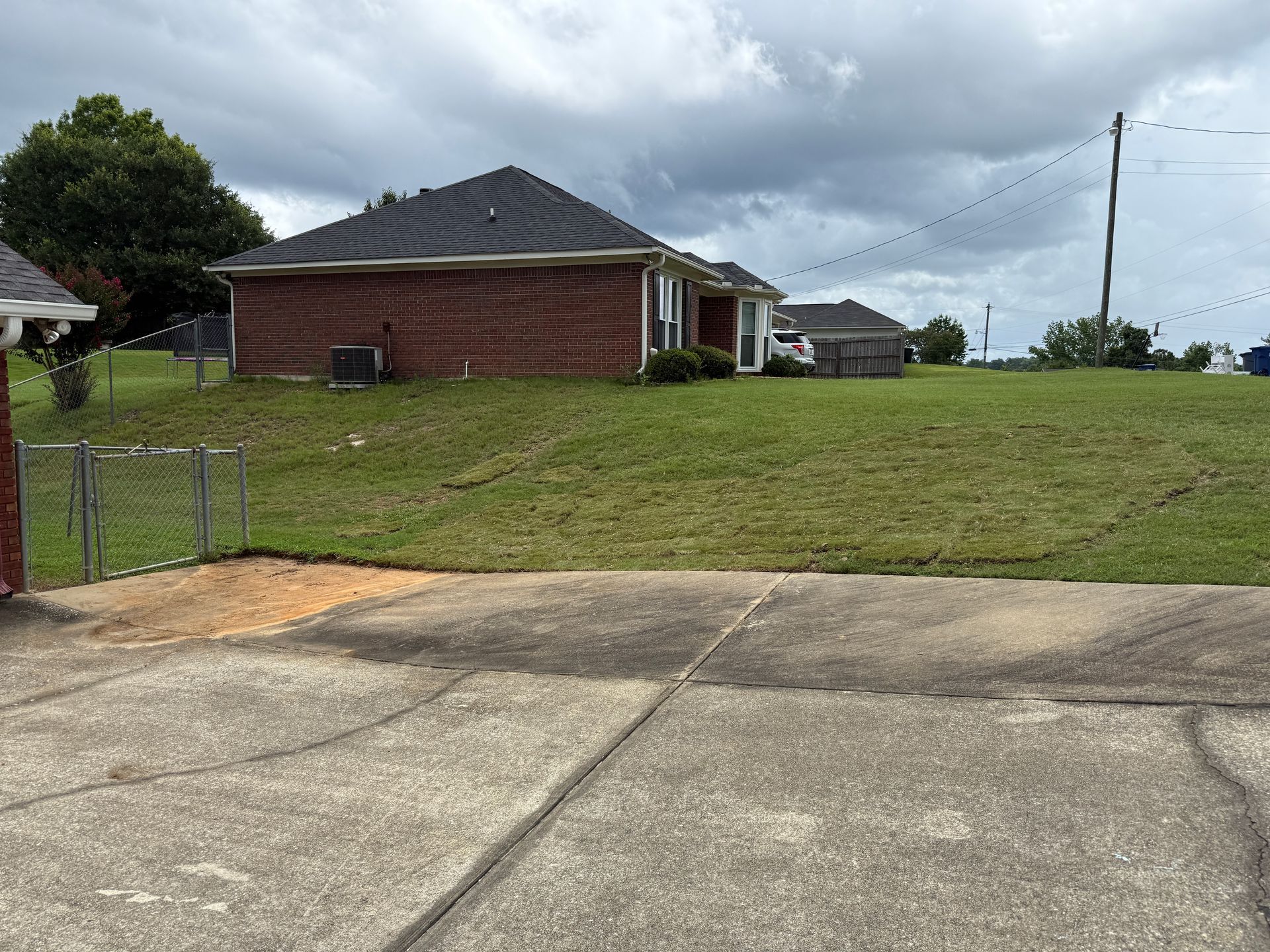 Brick house on a grassy hill; gray sky. Concrete in foreground, chain link fence at edge.