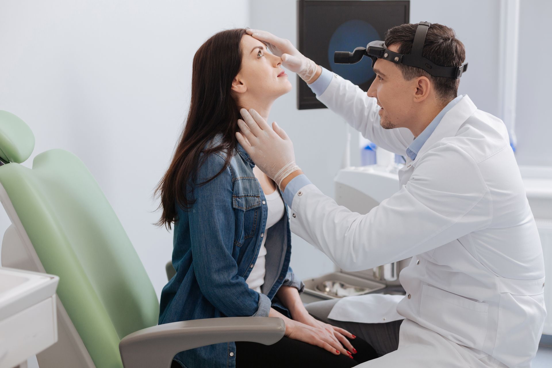 Doctor examining a seated patient’s throat in a clinic, with a chair and medical equipment nearby.