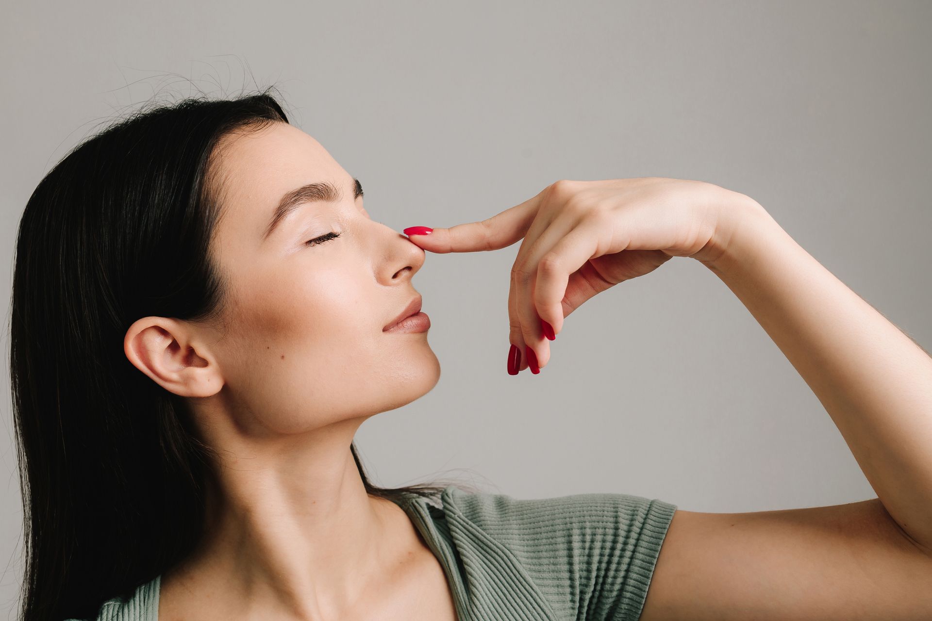 A person with dark hair closes their eyes and gently touches the tip of their nose with an index finger.