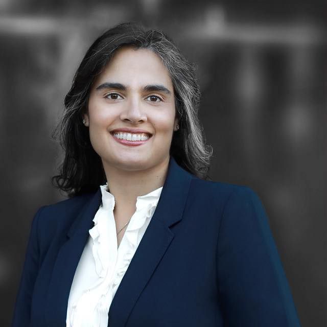 Woman with dark hair smiles, wearing a blue blazer and white ruffled blouse; blurred background.