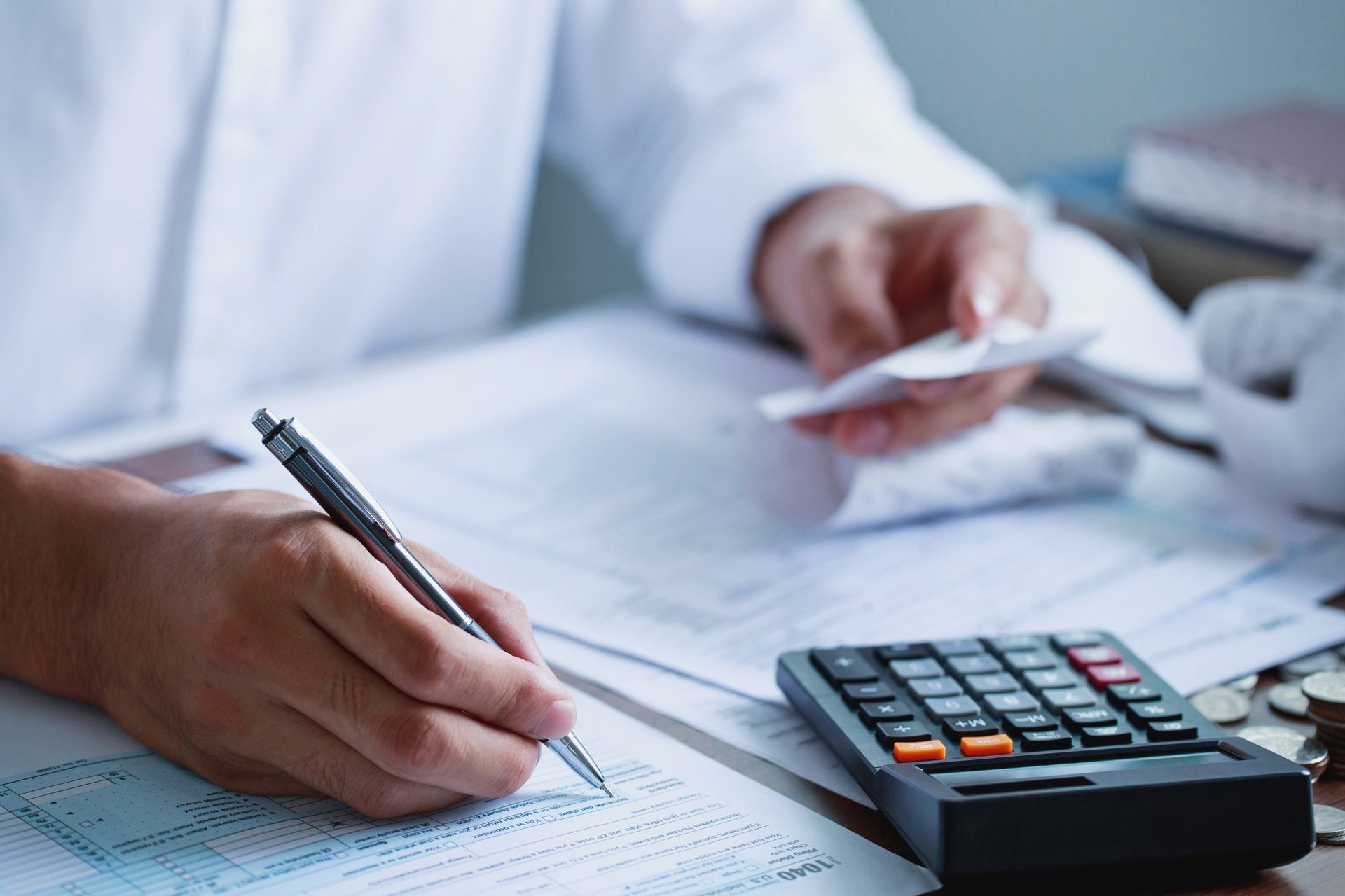 Person writing on paper with pen, holding receipts, using calculator and coins on desk.