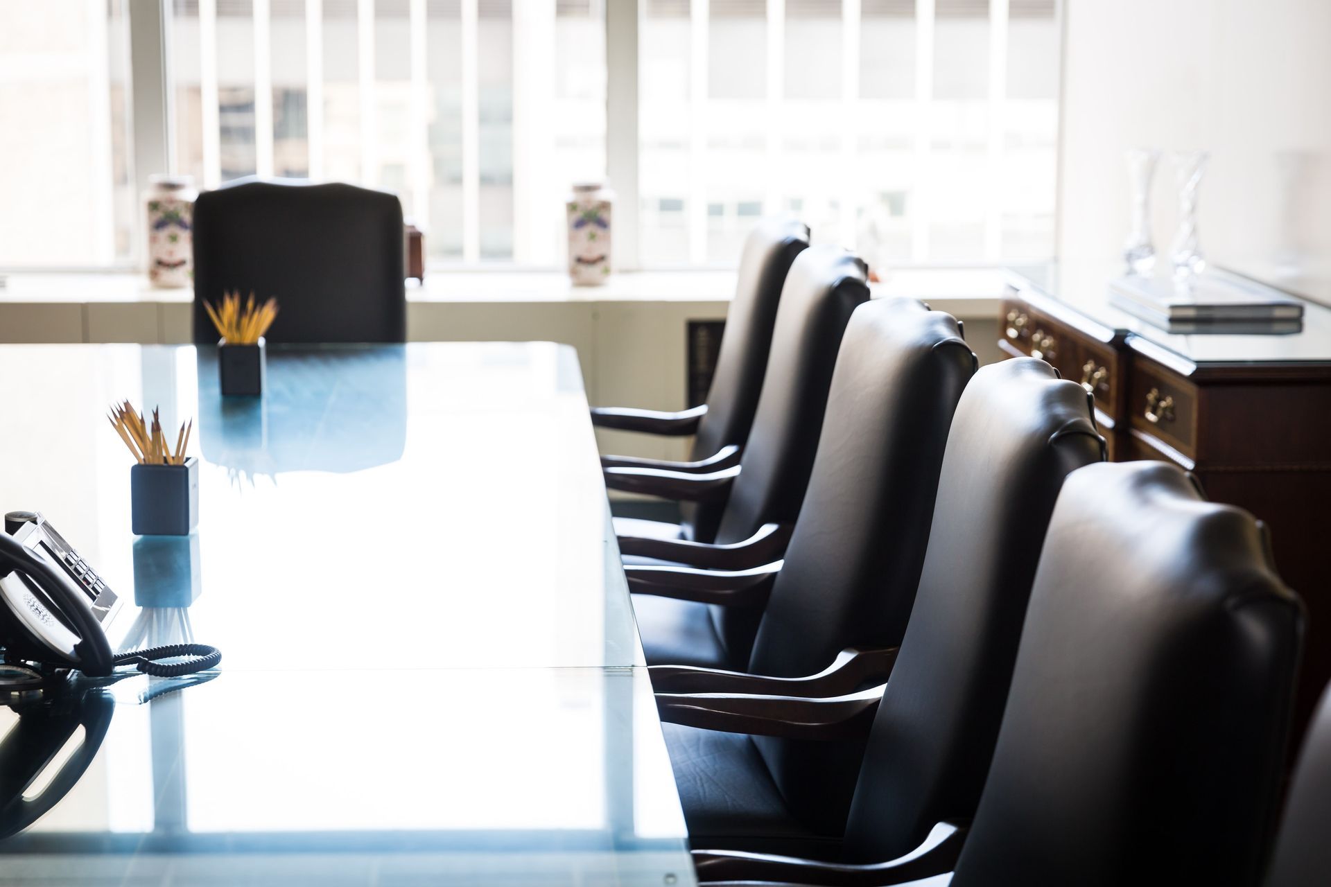 Conference room with a large, shiny table and black leather chairs. Sunlight streams in from a window.