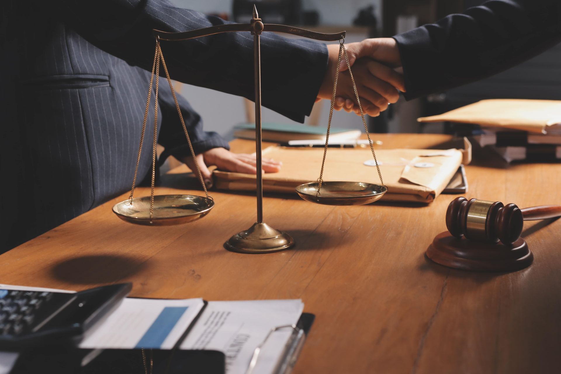 People shaking hands over a desk with scales, a gavel, and documents in a law office.