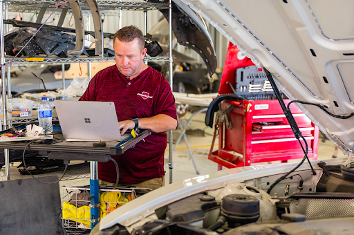 A man is using a laptop while working on a car in a garage.