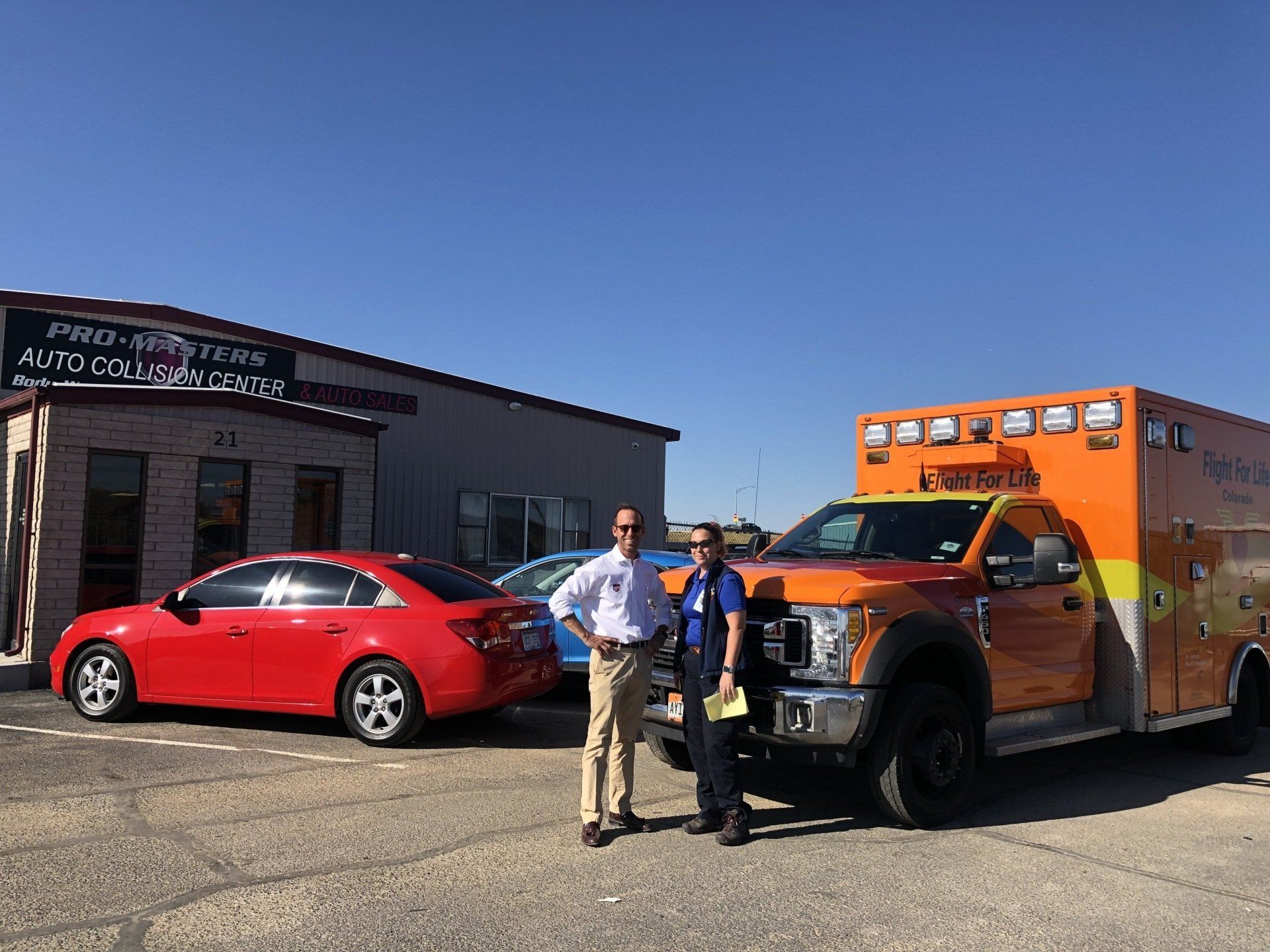 Two men are standing in front of an ambulance and a red car.