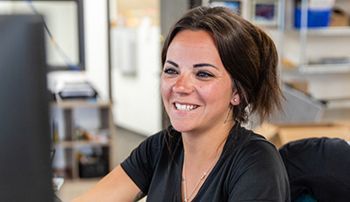 A woman is smiling while sitting at a desk in front of a computer.