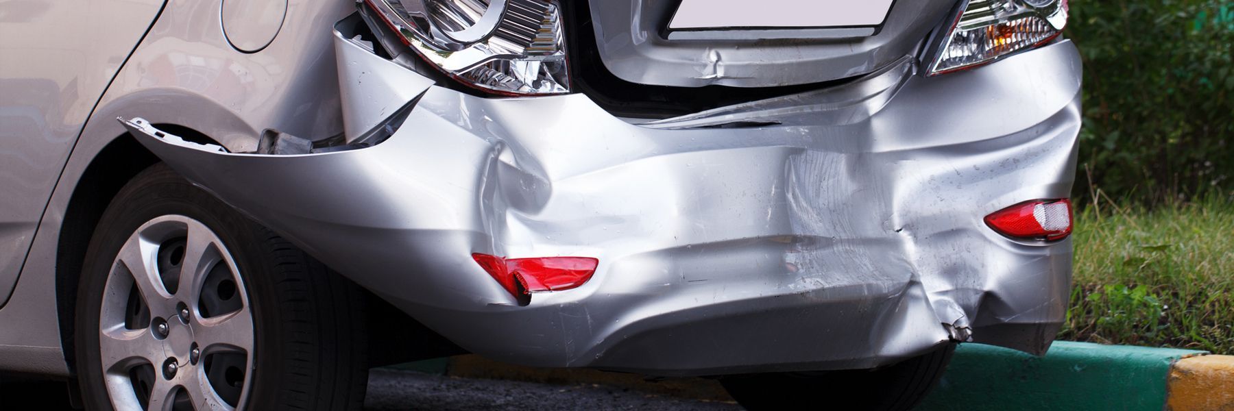 A silver car with a damaged bumper is parked on the side of the road.