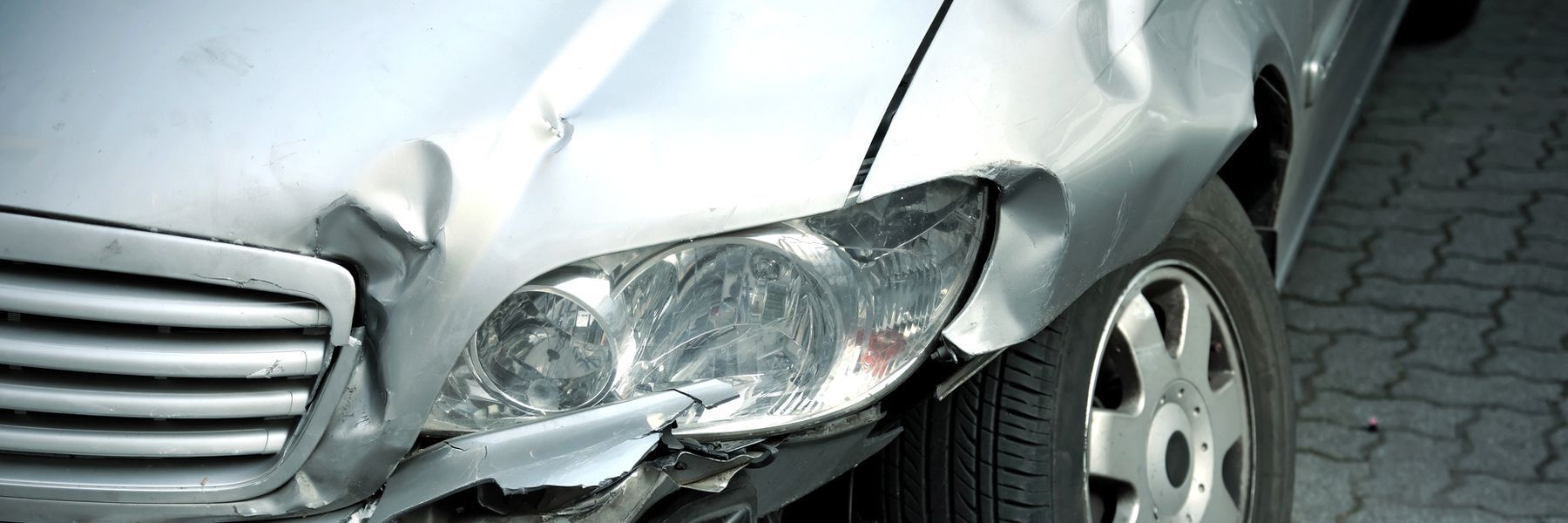 A silver car with a damaged front bumper is parked on a brick sidewalk.