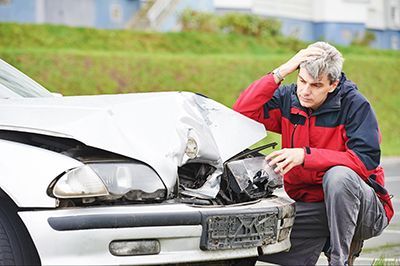 Man in red jacket kneels by car with damaged front, looking distressed outdoors.