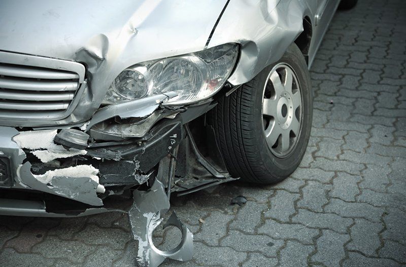 A silver car with a damaged front bumper is parked on a brick sidewalk.