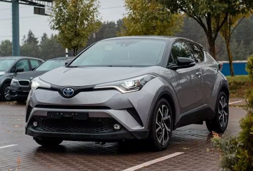 Gray Toyota C-HR hybrid parked on asphalt. Trees and other cars in the background.