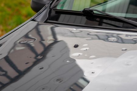 Hail-damaged gray car hood with numerous dents reflecting buildings and sky.