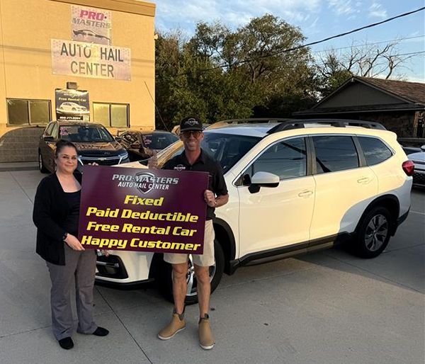 Two people standing next to a white SUV in front of an auto repair shop; holding a sign.