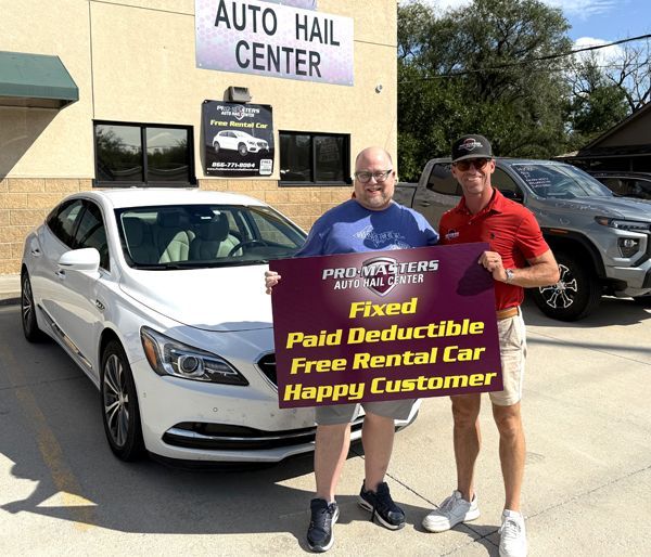 Two men smiling, holding a sign near a white car outside an auto hail center. The sign says