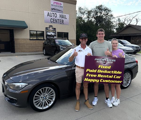 Man with two people in front of car at a business; they are holding a sign advertising auto hail services.