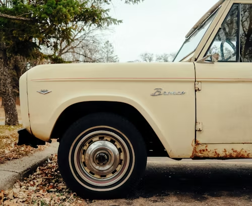 Tan vintage Ford Bronco with rusty detailing, parked near a tree on a fall day.