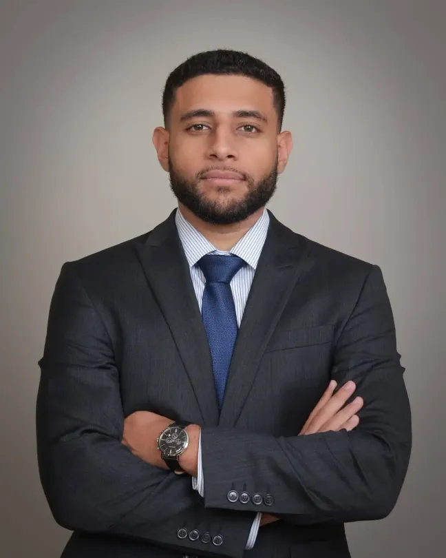 Portrait of a man in a dark suit and blue tie, standing with arms crossed against a gray background