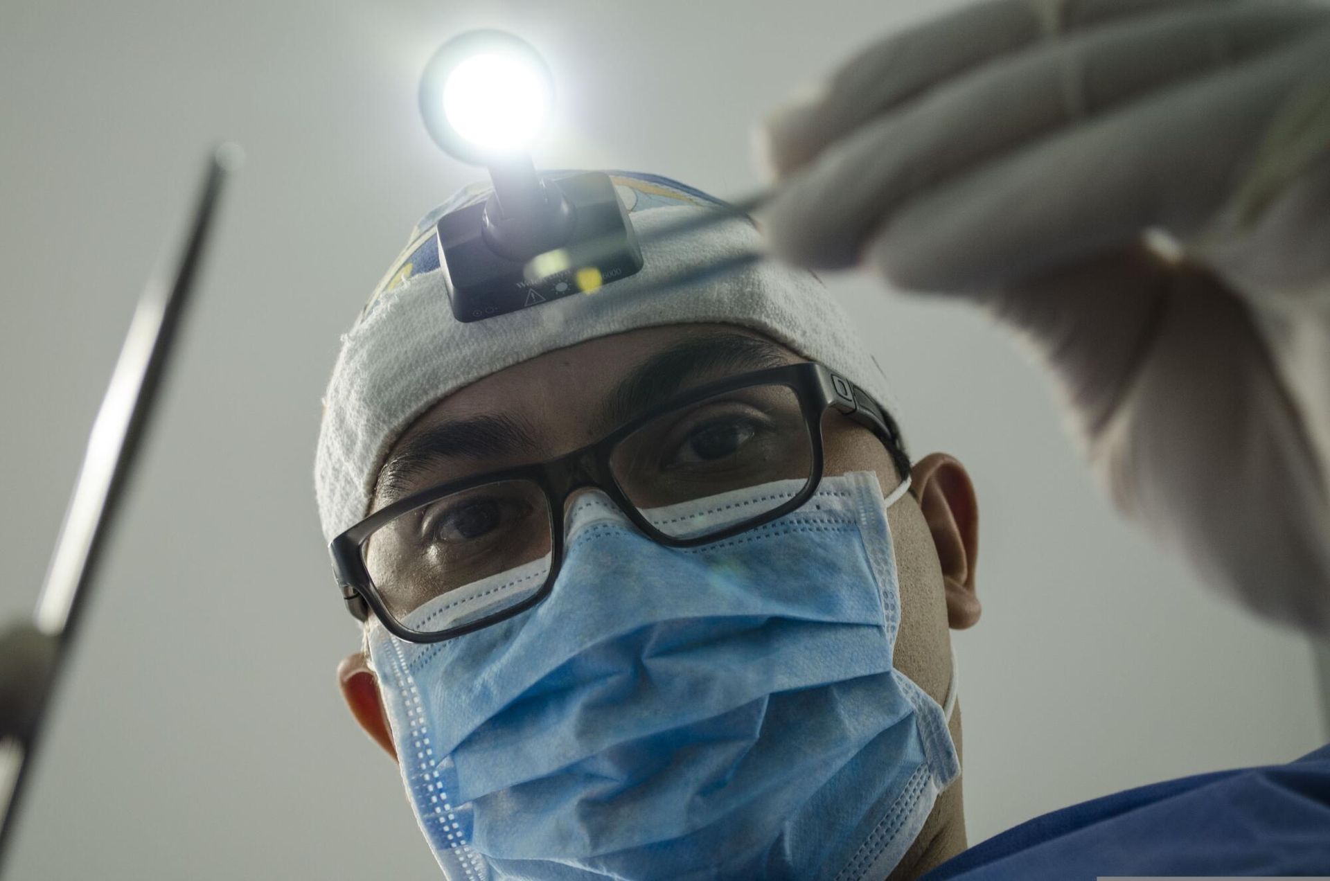 Dentist leaning over patient, holding tools, wearing mask, glasses, headlamp.