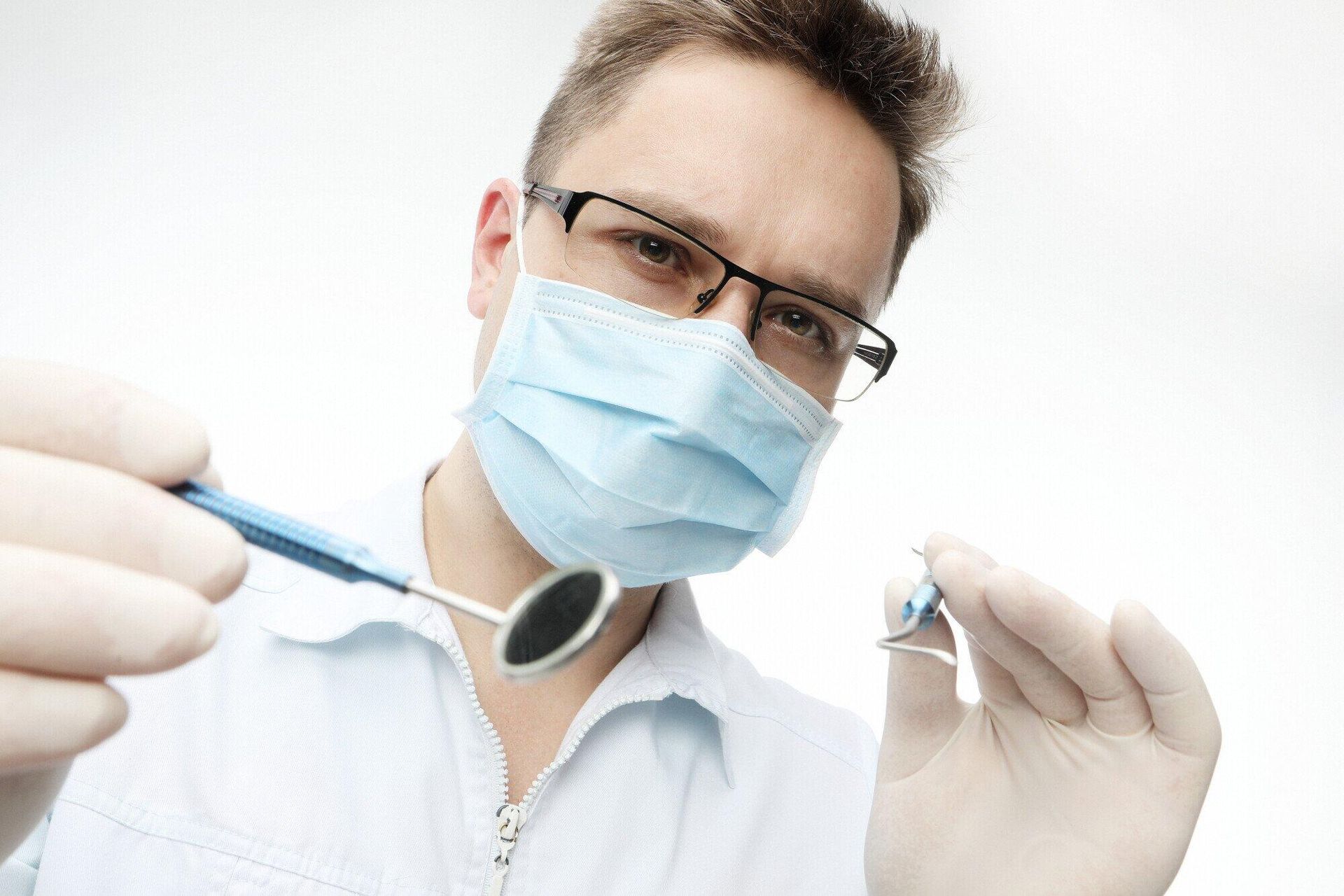 Dentist wearing mask and glasses, holding dental tools, looking down at patient.