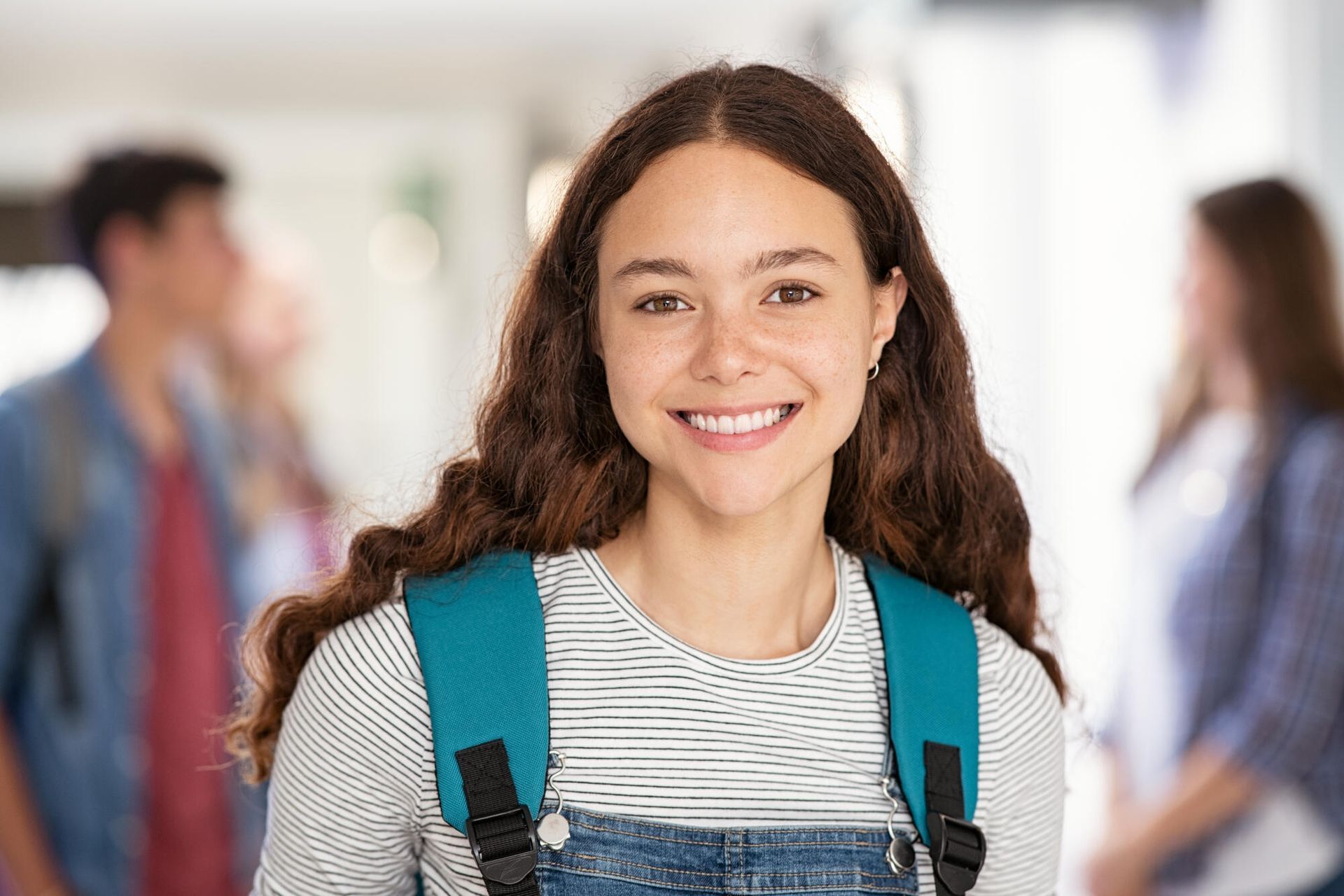 Smiling student with brown wavy hair and a teal backpack in a school hallway.