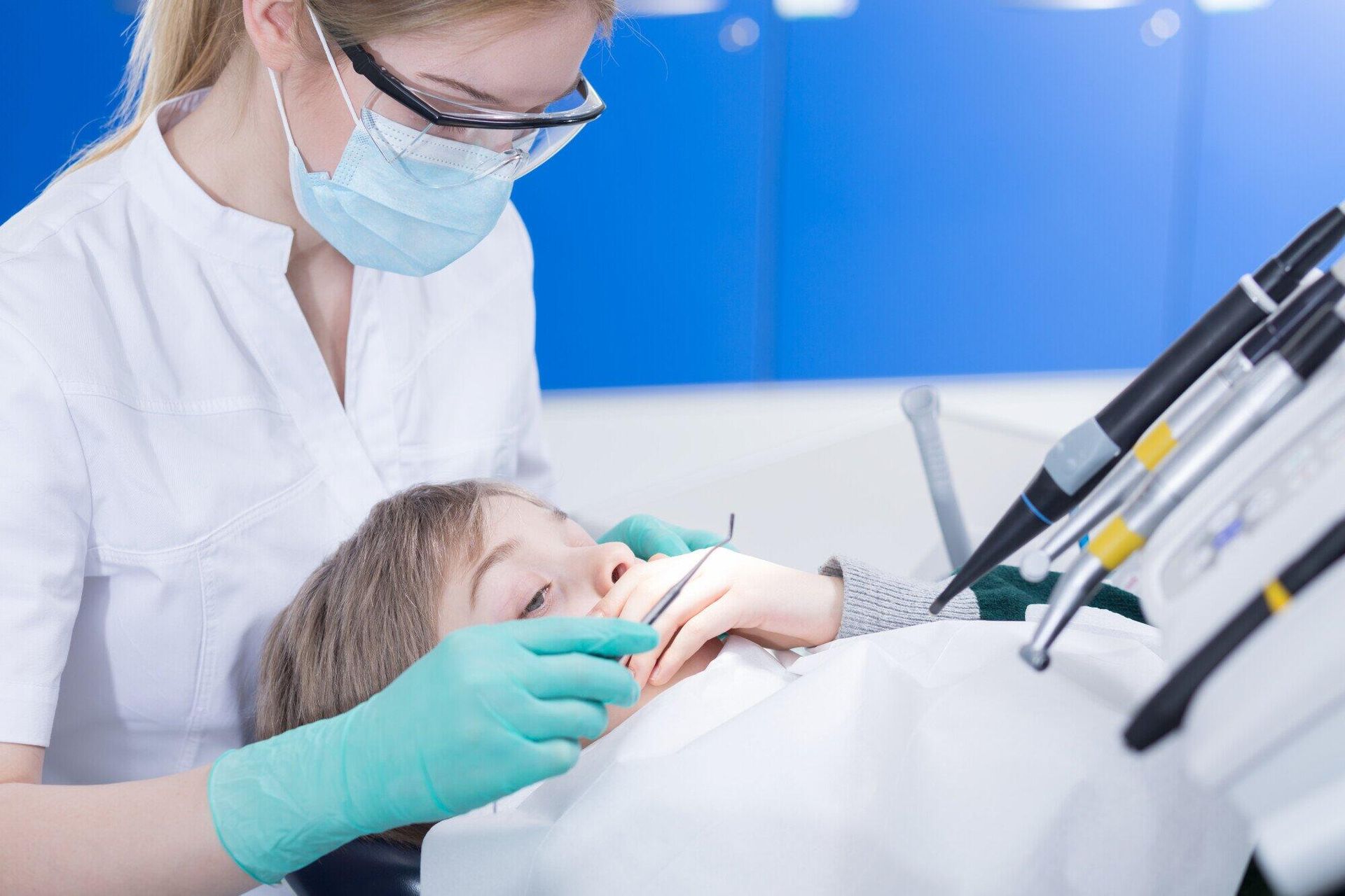 Dentist in protective gear examining a child's teeth in a dental chair.