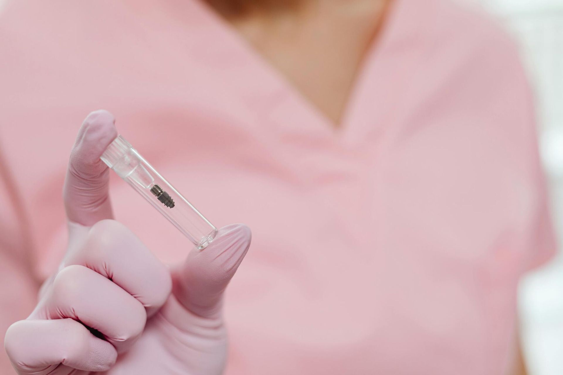 Person in pink scrubs and gloves holding a small vial with a dark object inside.