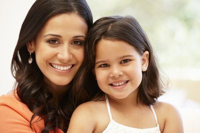 Woman with dark hair smiles with a young girl. They are close together and appear happy.