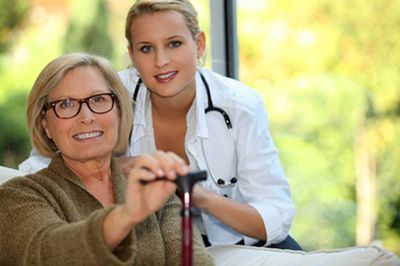 Woman with cane smiles, supported by a caregiver indoors with stethoscope.