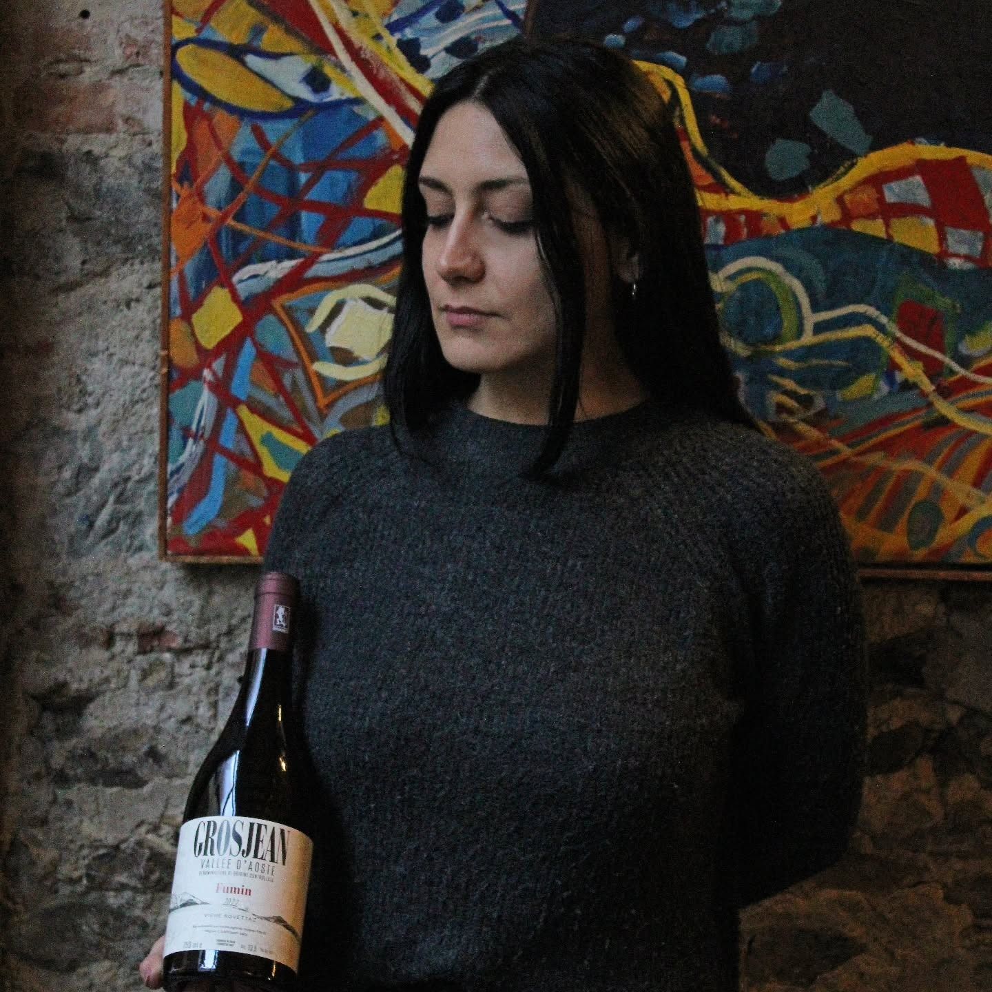 Donna con in mano una bottiglia di vino, guardando in basso. In una stanza con pareti in mattoni e un dipinto colorato sullo sfondo.