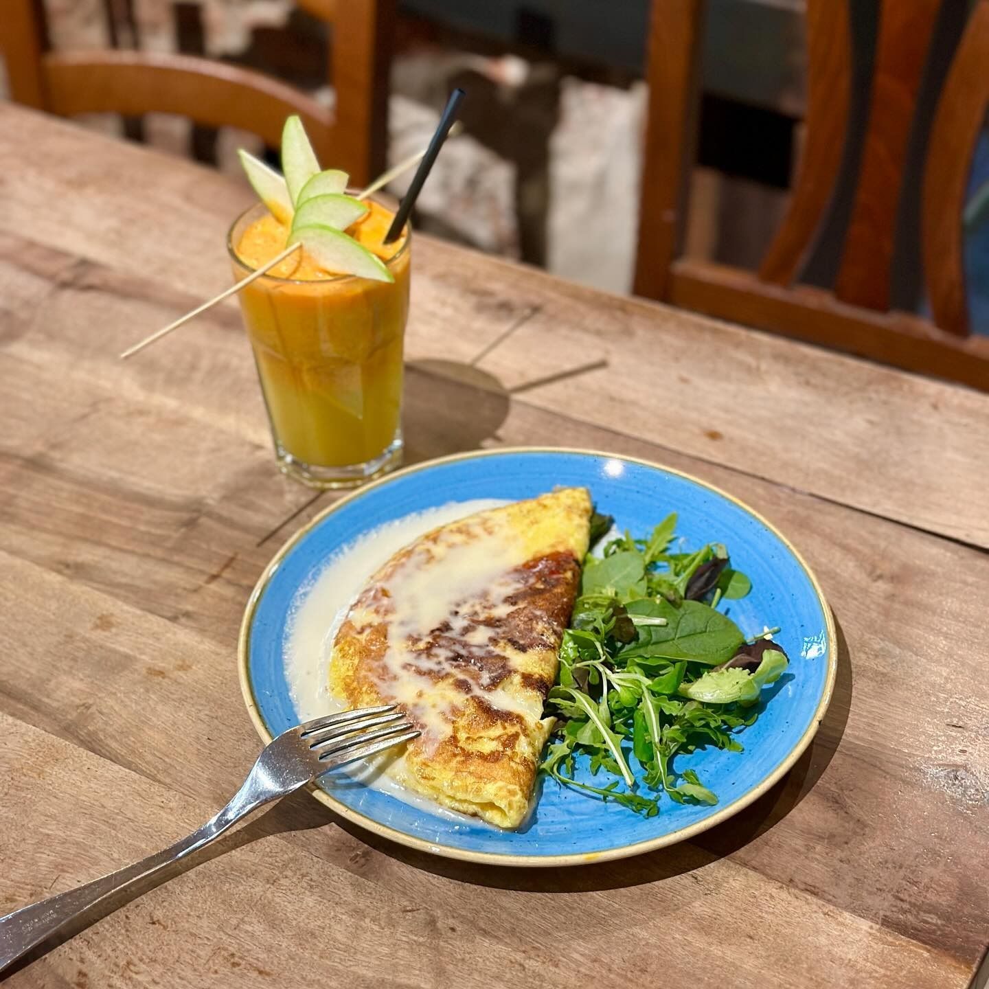Frittata e insalata su un piatto blu, con una bevanda color carota guarnita con fette di mela su un tavolo di legno.