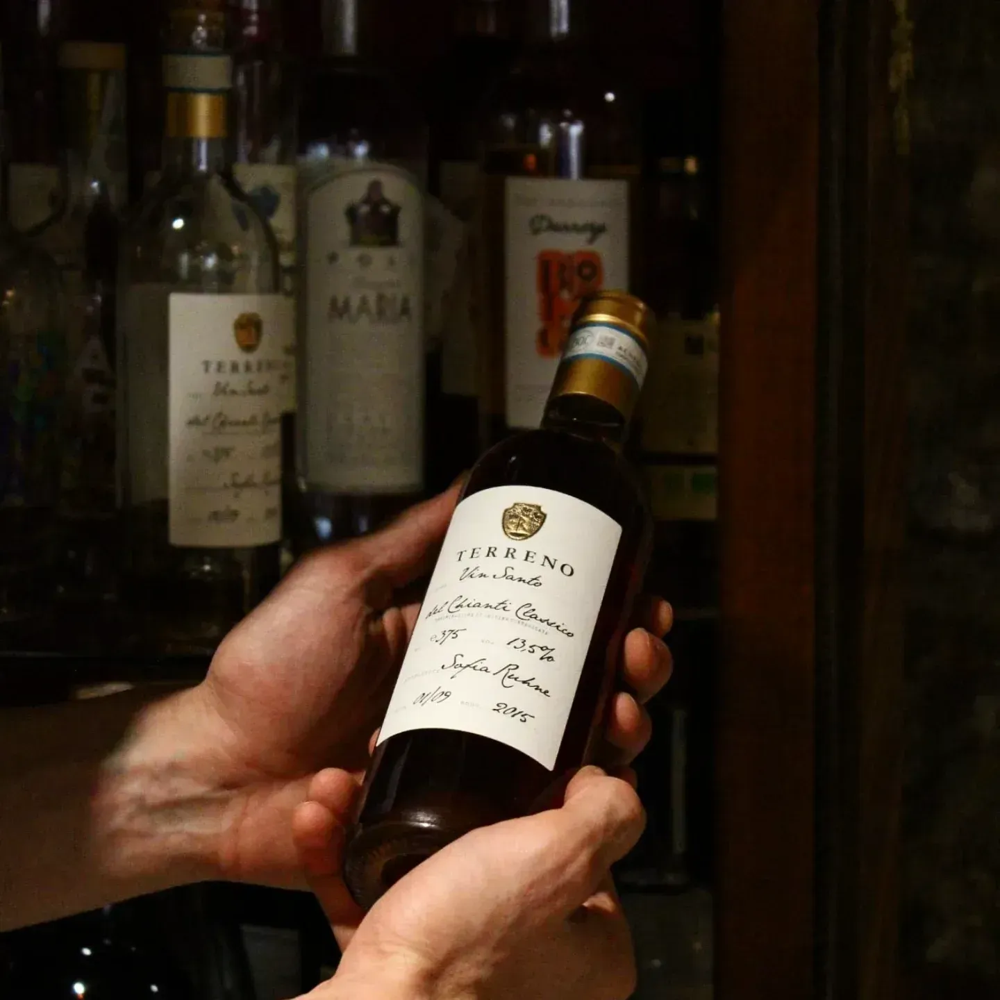 Mani che tengono una bottiglia di vino davanti a uno scaffale pieno di altre bottiglie.