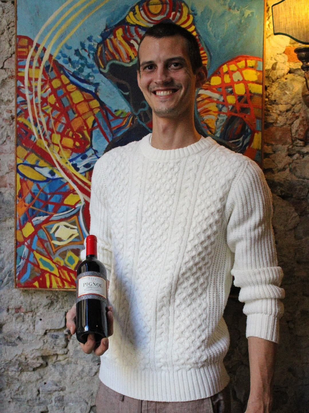 Uomo che tiene in mano una bottiglia di vino rosso e sorride davanti a un dipinto astratto colorato.