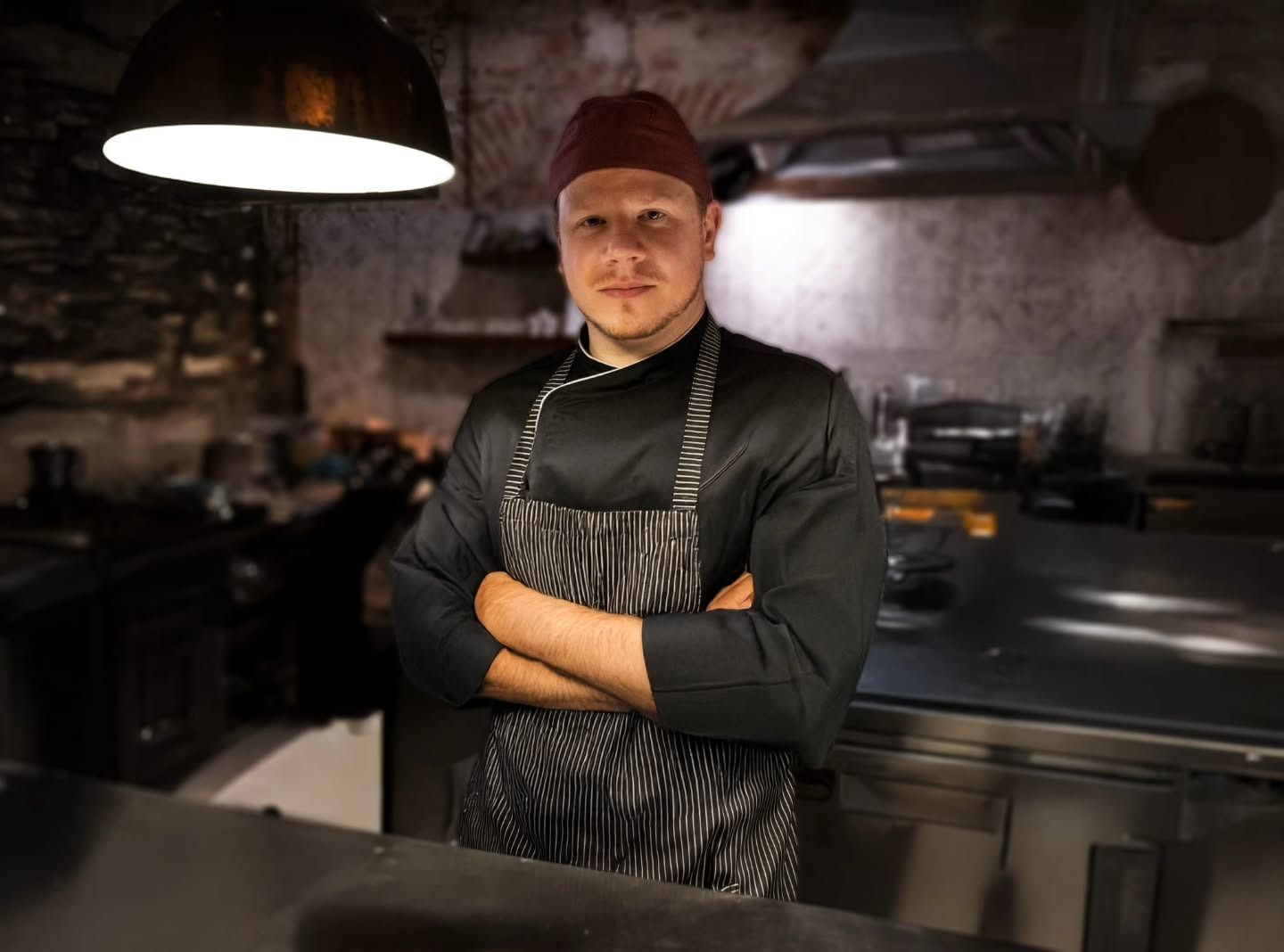 Cuoco in uniforme nera e grembiule a righe, braccia incrociate, in una cucina con luce dall'alto.