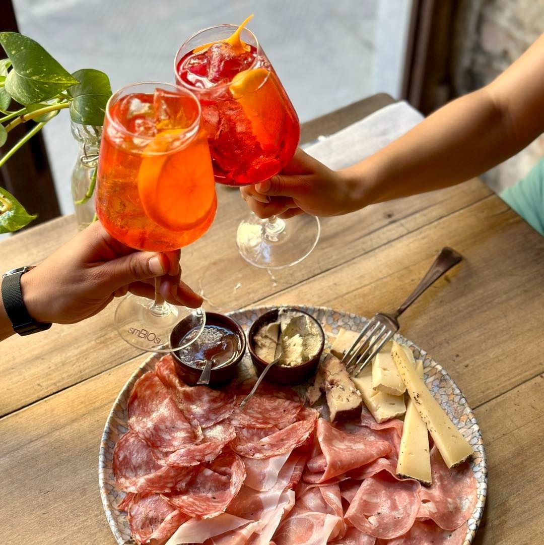 Due persone che brindano con gli spritz di Aperol su un tagliere di salumi, formaggi e salse, su un tavolo di legno.