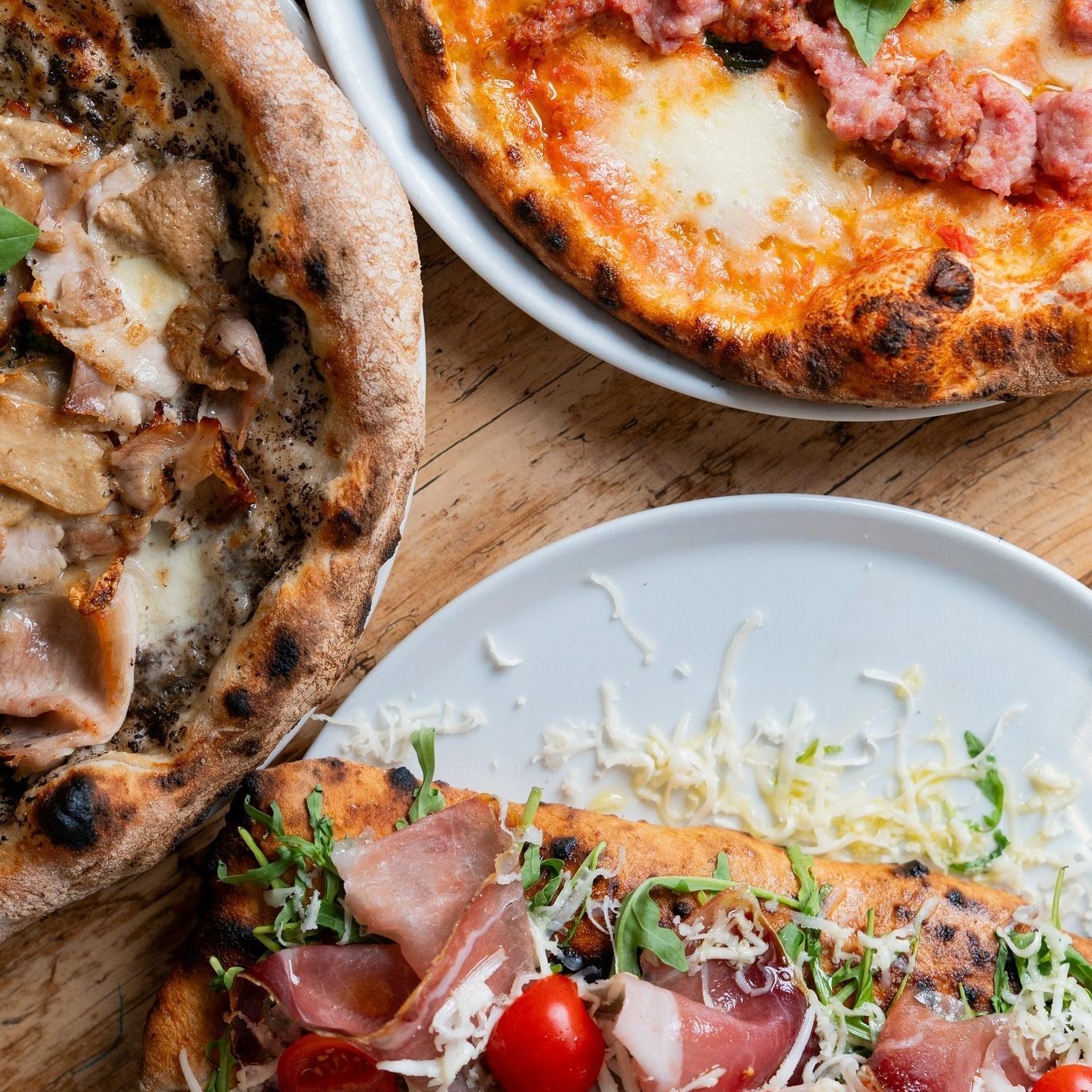 Tre pizze su superficie di legno: funghi, salsiccia e prosciutto con rucola, pomodori e formaggio.